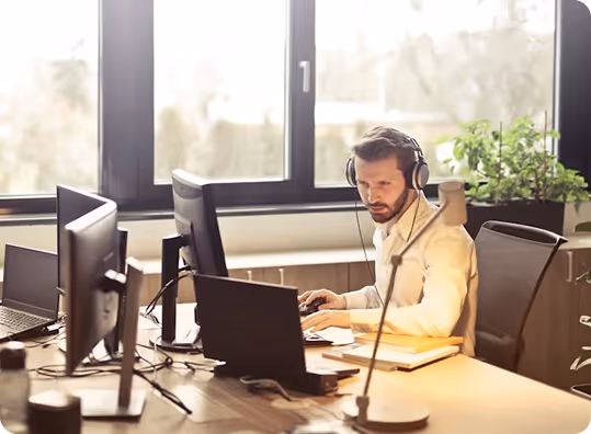 Man wearing headphones working on laptop at desk near large windows in bright office.