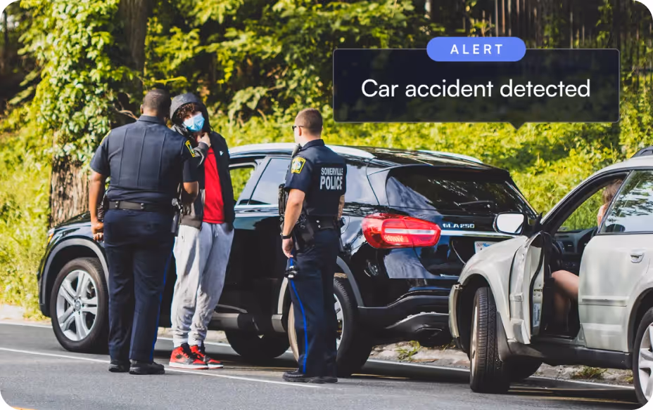 Two police officers question a masked man near vehicles after a detected car accident alert.
