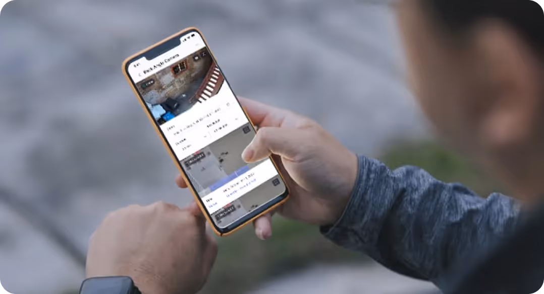 Person holding a smartphone displaying multiple security camera live feeds outdoors.