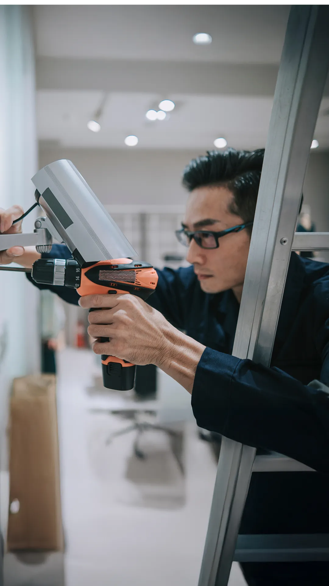 Man wearing glasses installs a security camera using an orange cordless power drill on a ladder.