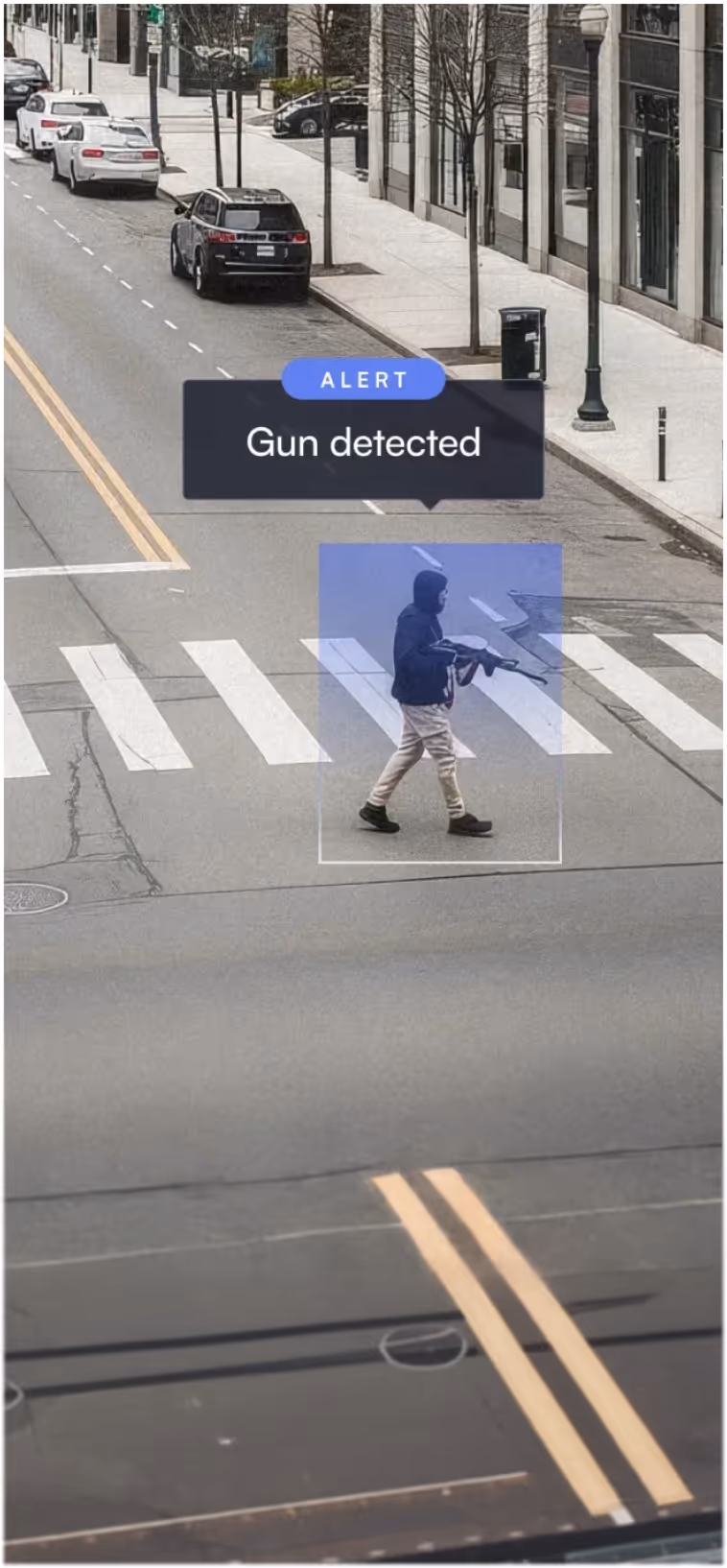 Surveillance image showing a person crossing the street holding a firearm with alert text reading Gun detected.