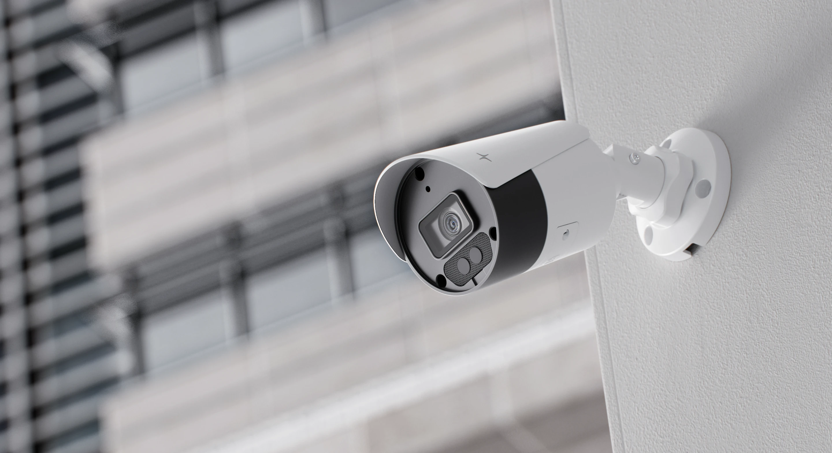 White security camera mounted on a textured white wall with blurred building background.