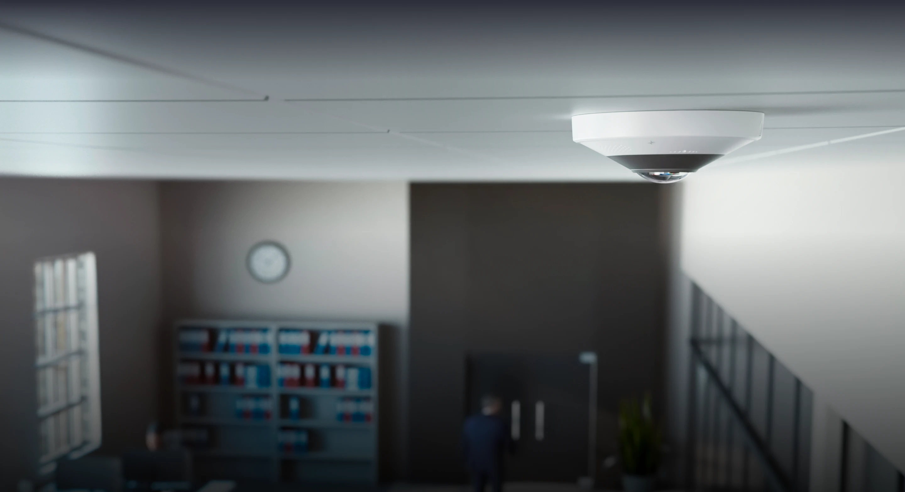 White dome security camera mounted on ceiling in an office with shelves, a clock, and a person near doors.