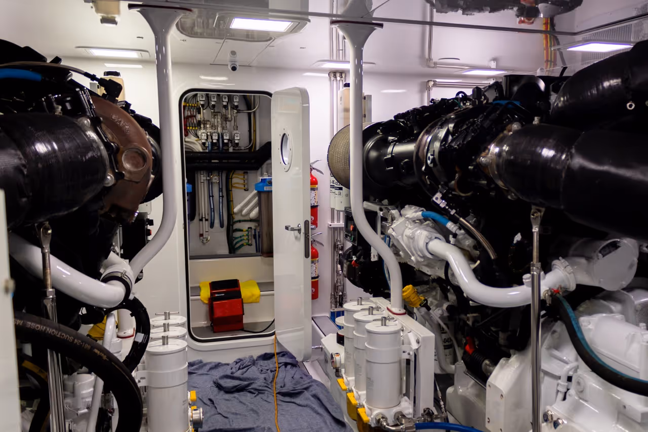 Interior of a boat engine room showing two large engines, piping, fire extinguishers, and an open door leading to a control panel.