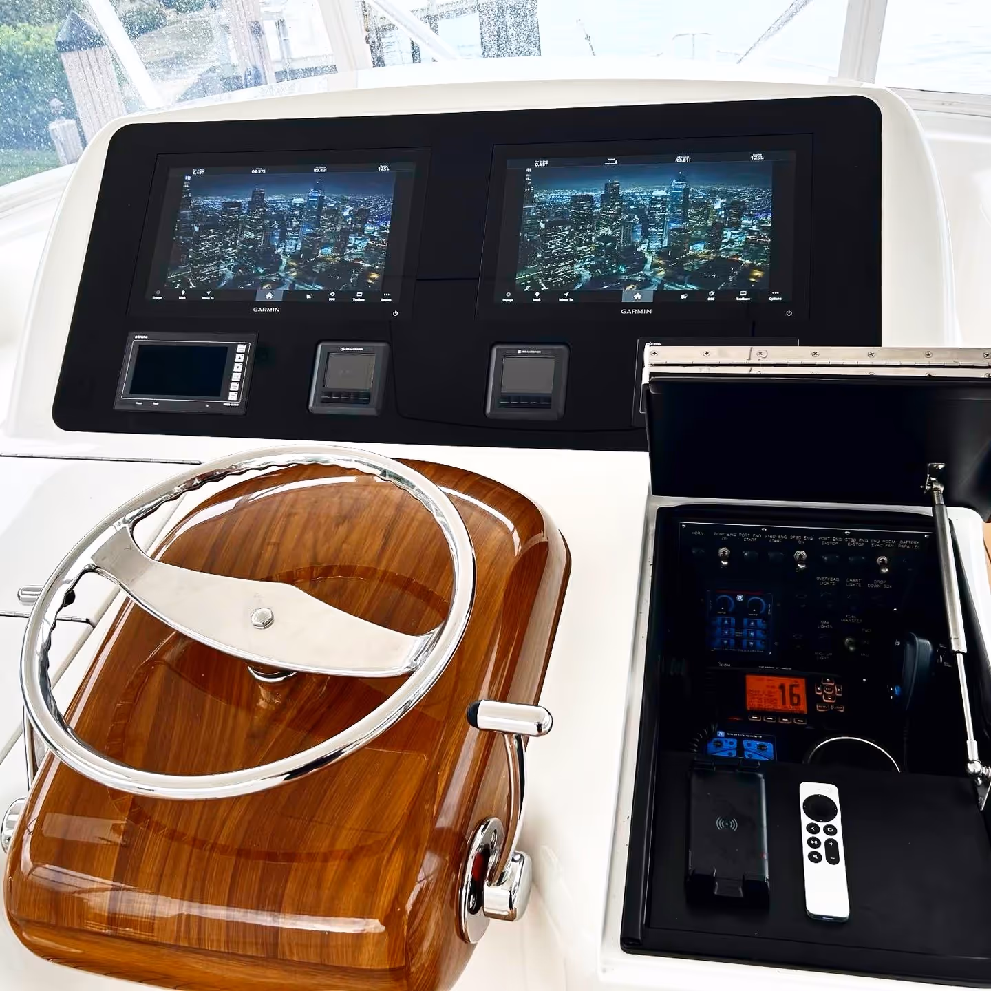 Boat steering console with wooden helm, dual Garmin navigation screens, control switches, and an open compartment containing communication and remote control devices.