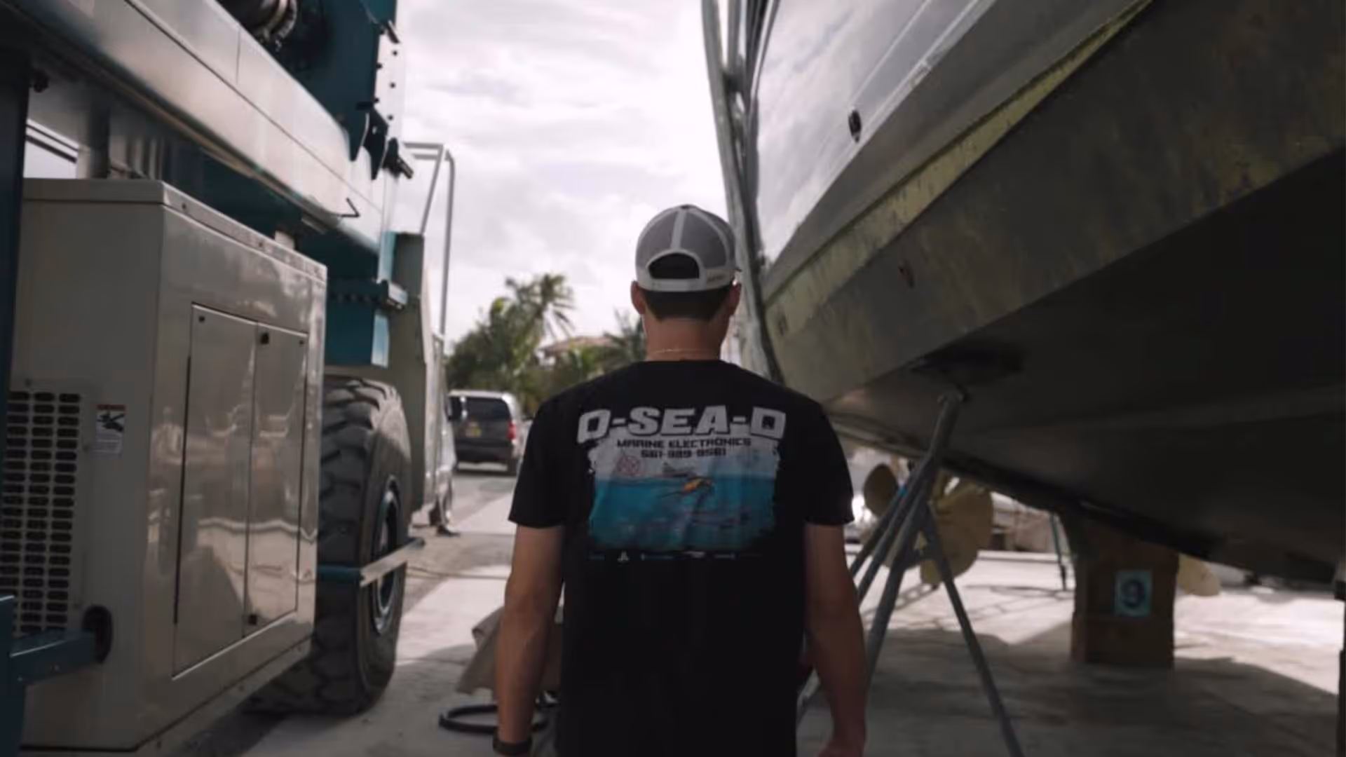 Man wearing a black T-shirt and gray cap walking beside a large boat on supports.