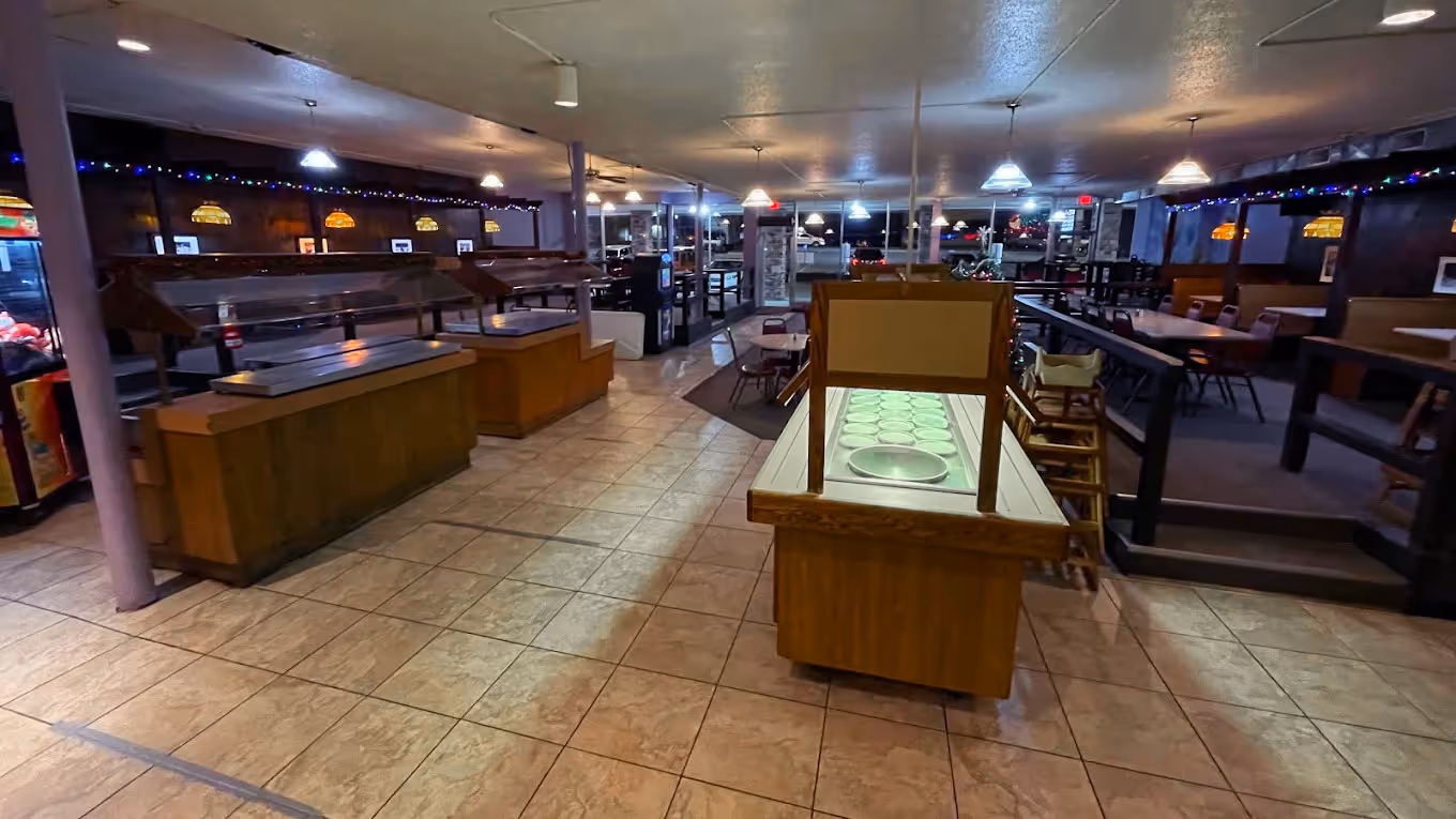 A spacious, dimly-lit restaurant interior with a tiled floor, buffet counters, and empty dining tables.