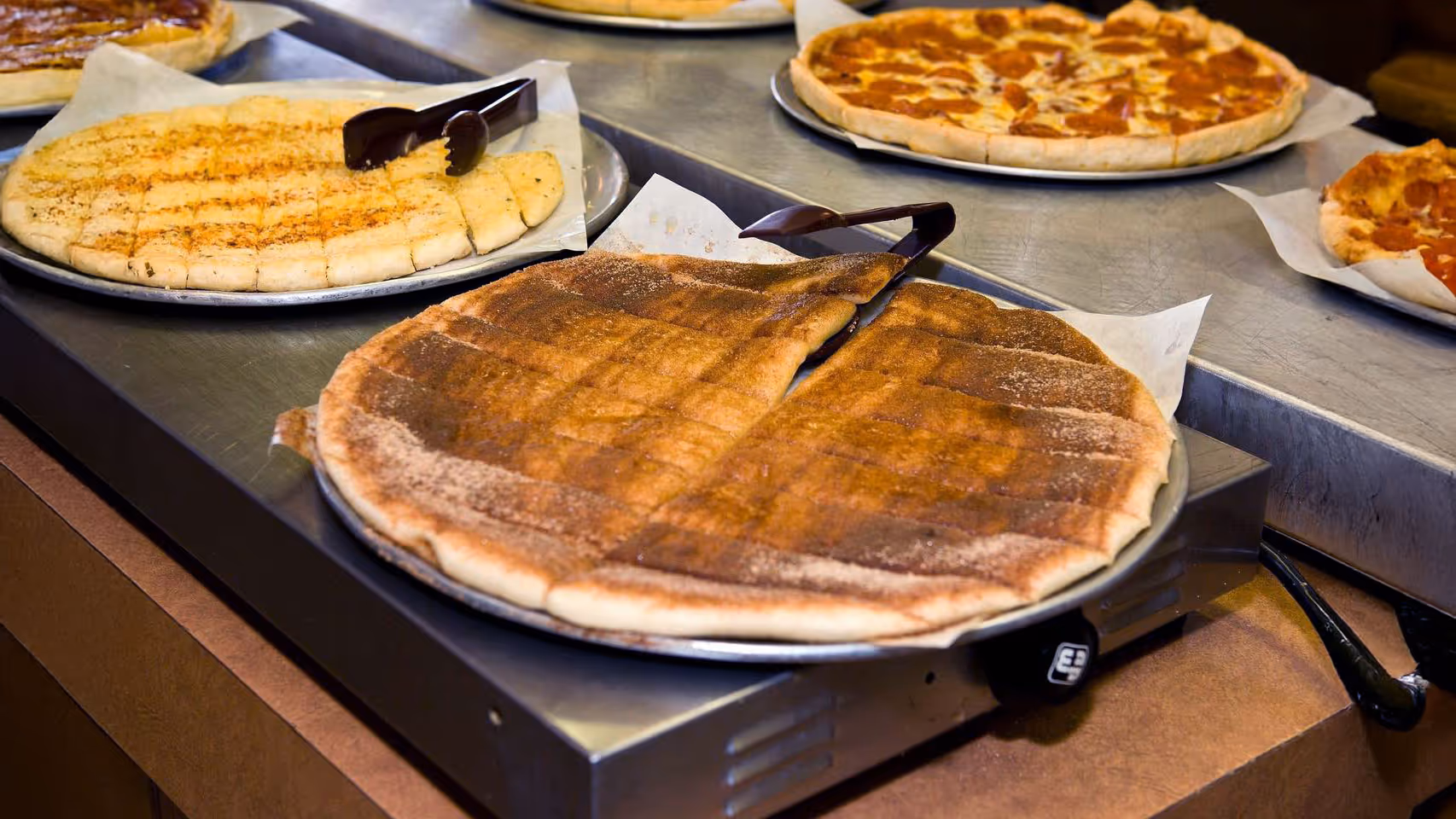 An assortment of pizzas on metal trays: a cinnamon dessert pizza with a serving spatula, a cheesy pizza, and a pizza with a grid pattern, all on a buffet table.
