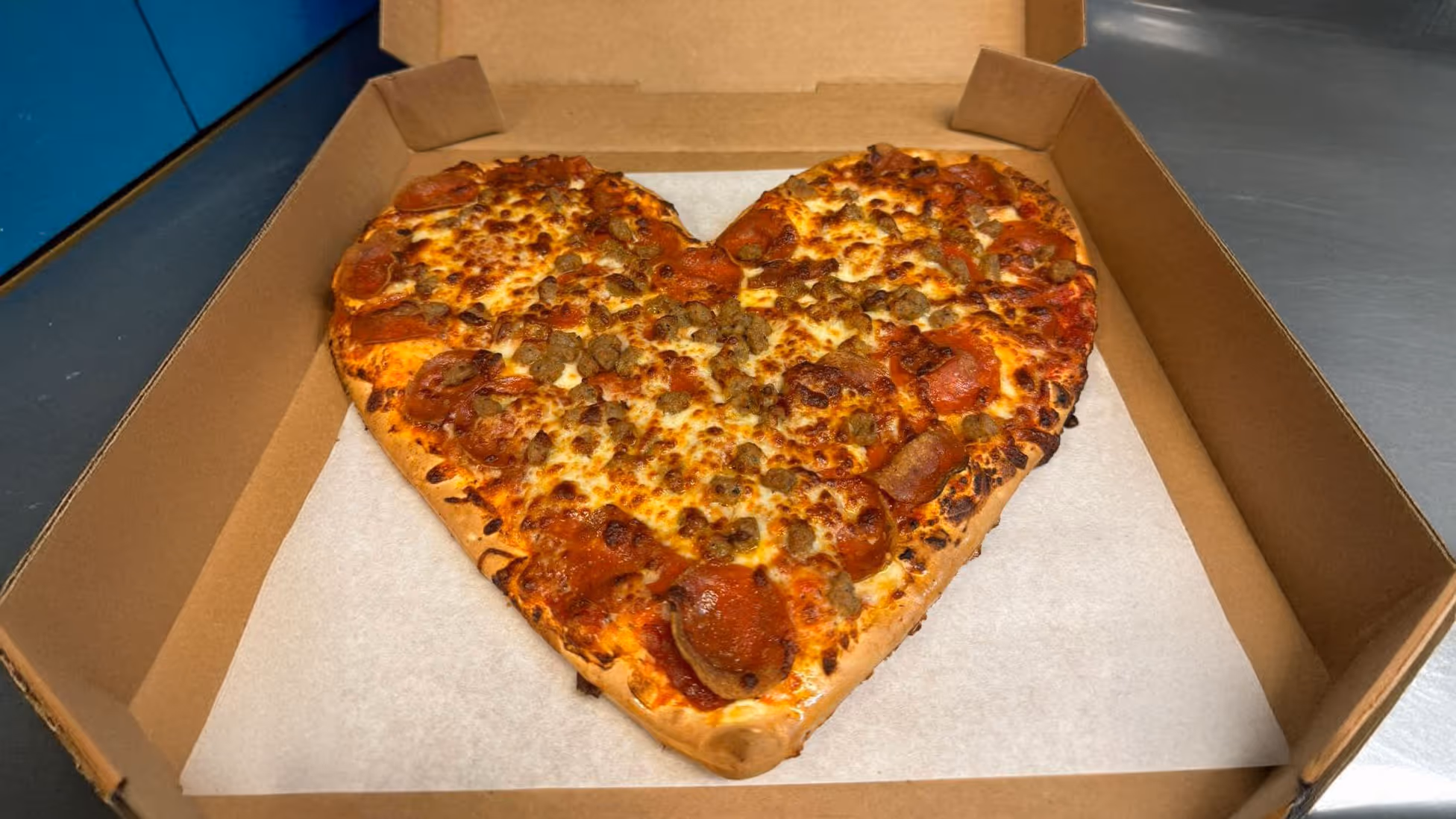 A heart-shaped pizza with golden crust, topped with pepperoni and sausage, in an open cardboard box on a metal counter.