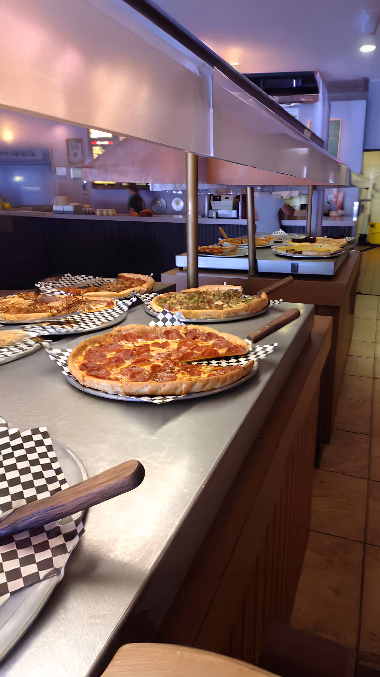 A buffet lineup featuring various pizzas, each on checkered paper atop a silver counter. The scene conveys a casual, inviting dining atmosphere.