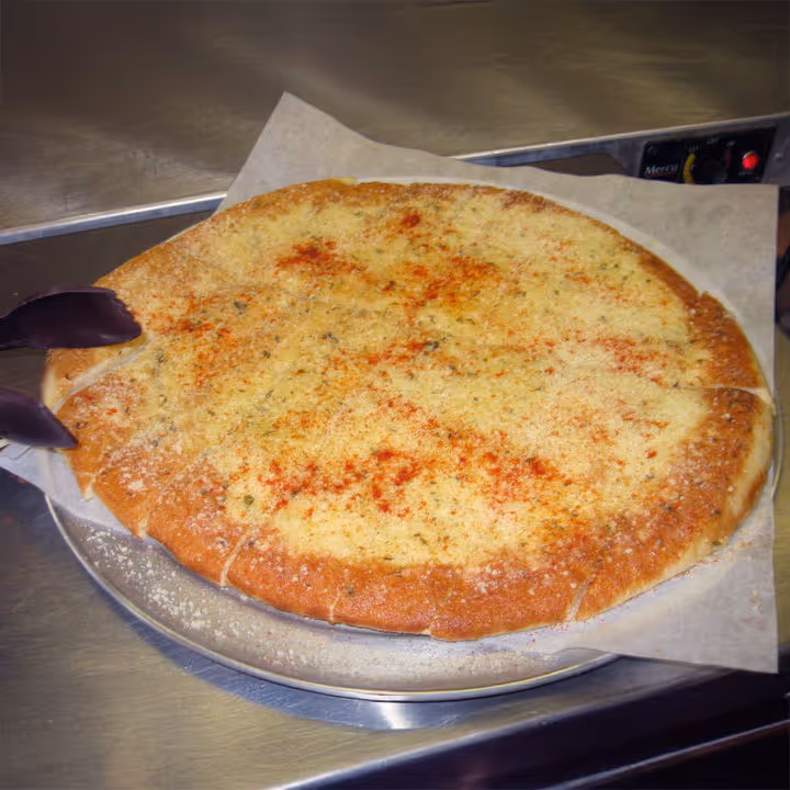 A large cheese-covered pizza on a metal tray, topped with red seasoning and cut into slices. It rests on parchment paper in an industrial kitchen setting.
