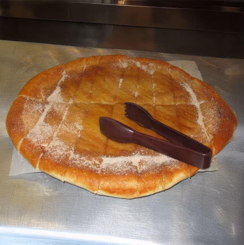 A large cinnamon and sugar-coated pastry flatbread sliced into squares, with plastic serving tongs on top, on a metal countertop.