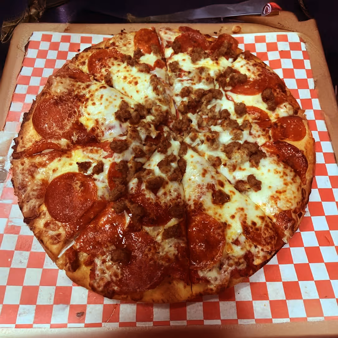 A pizza with a generous topping of pepperoni and sausage, on a checkered red and white paper.