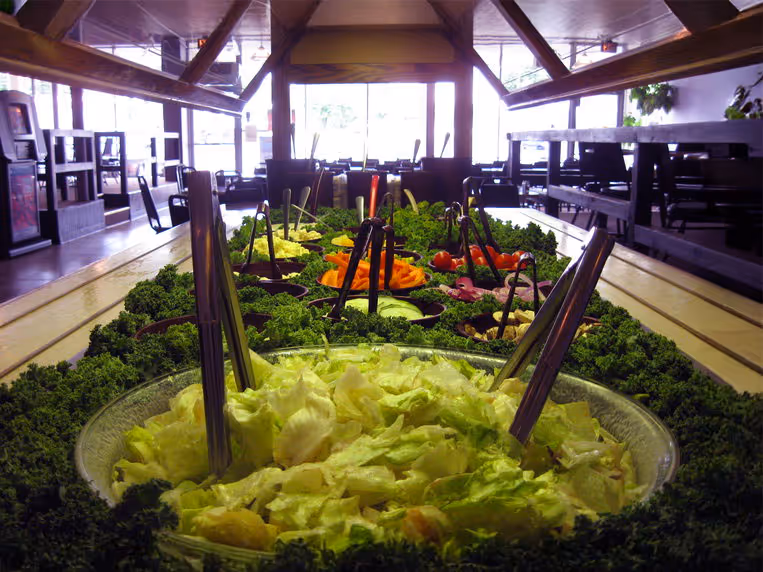 A salad bar with fresh lettuce, colorful vegetables, and toppings arranged in bowls on a long counter.