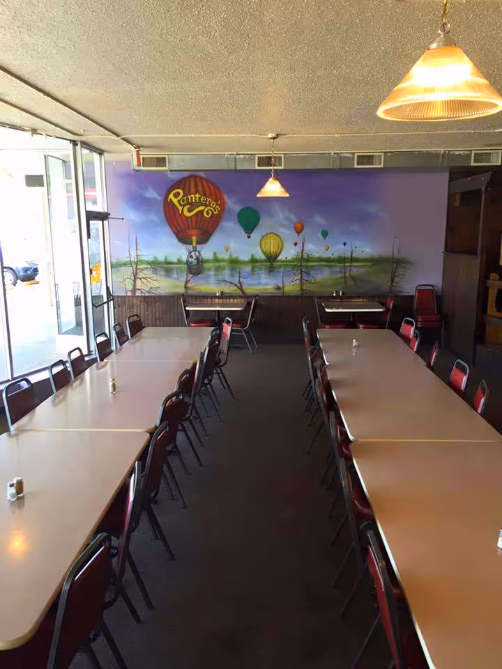 A restaurant interior with long tables and red chairs. A mural of colorful hot air balloons and a landscape adorns the wall.