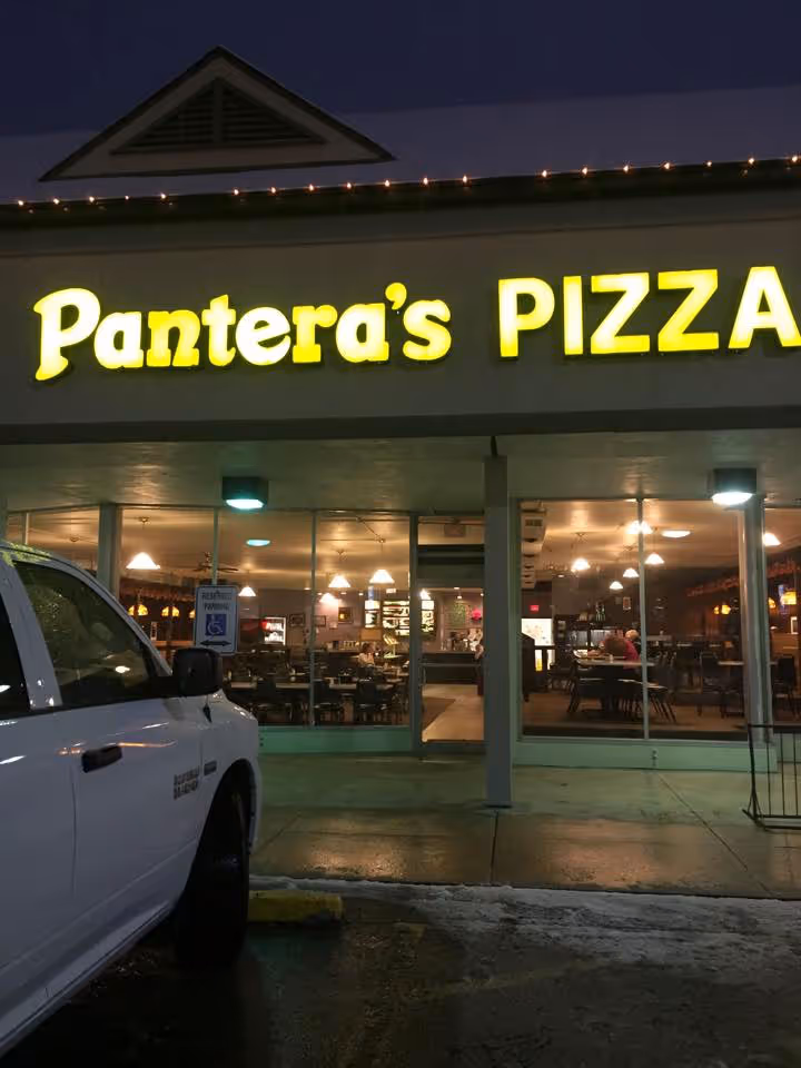 A nighttime view of Pantera's Pizza with lit yellow signage. Inside, warmly lit tables and chairs. A white pickup truck is parked outside. Cozy ambiance.