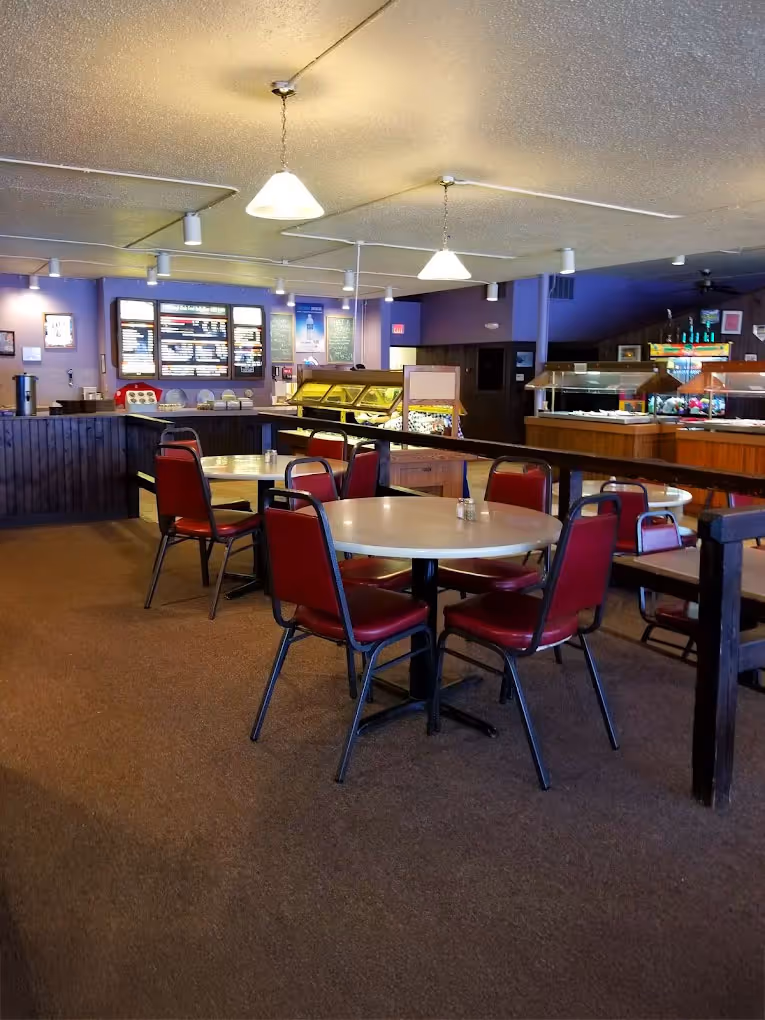 A cozy restaurant interior with round tables and red-cushioned chairs. Soft lighting and a visible counter with menu boards create a warm, inviting atmosphere.
