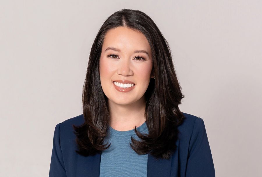 Smiling woman with shoulder-length black hair wearing a beige blazer against a gray textured background.