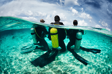Two scuba divers with oxygen tanks kneeling underwater on a clear sandy seabed beneath a partly cloudy sky.