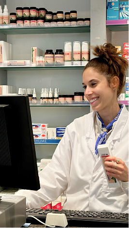 Smiling pharmacist working at computer with shelves of medicine behind