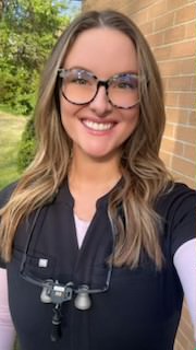 Smiling woman with long hair wearing tortoiseshell glasses and black scrubs, standing outdoors near a brick wall.