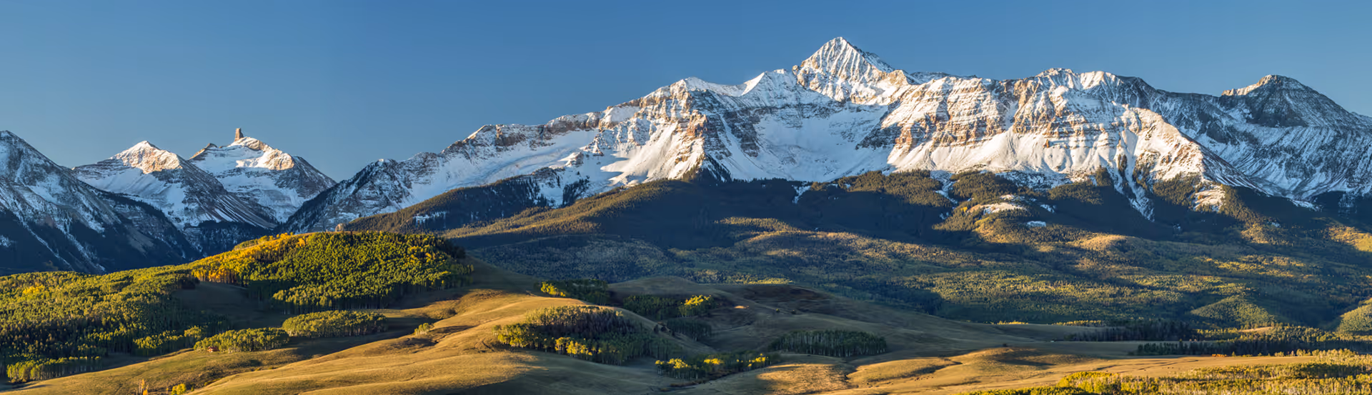 Snow-covered mountain range with a clear blue sky and sunlit green and brown foothills.