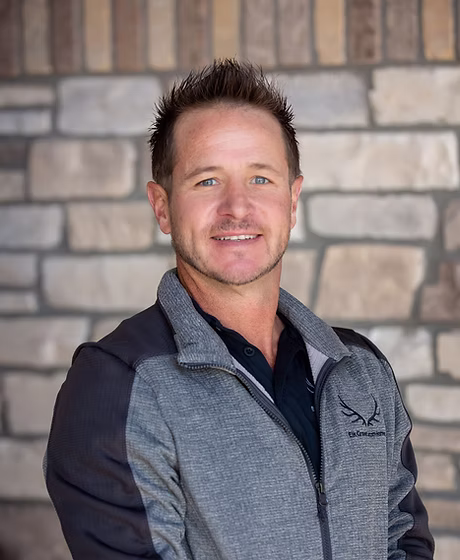Smiling man with short spiky hair wearing a gray and black zip-up jacket standing in front of a stone wall.
