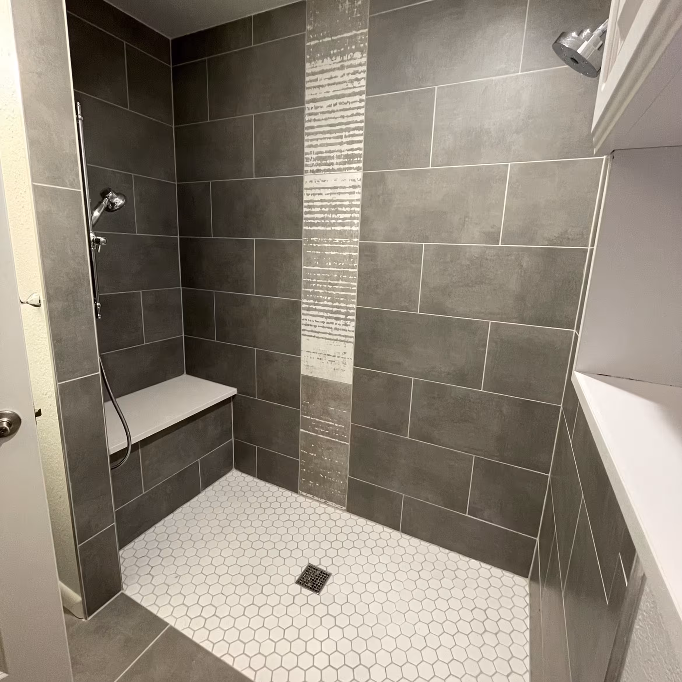 A modern walk-in shower features large, dark gray wall tiles accented by a vertical strip of textured, lighter gray decorative tile. The floor is composed of white hexagonal mosaic tiles, sloping toward a central square drain for easy drainage. Inside, a built-in white shower bench is positioned beneath a handheld showerhead, while a second rain-style showerhead is mounted on the opposite wall.