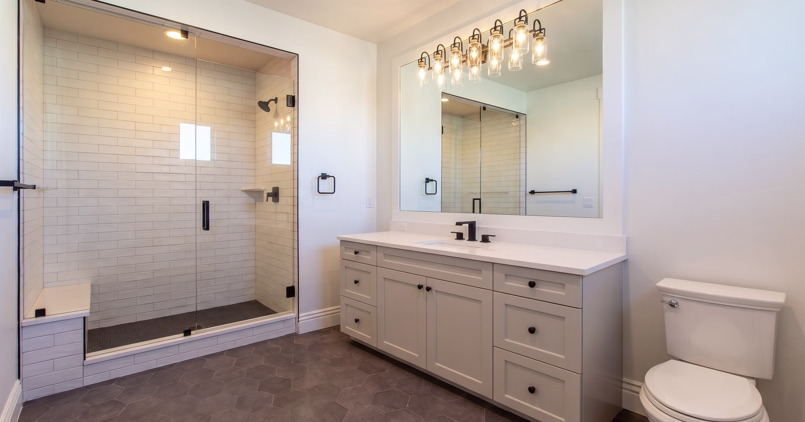 Modern bathroom with glass shower, white vanity with black fixtures, and white toilet on gray hex tile floor.