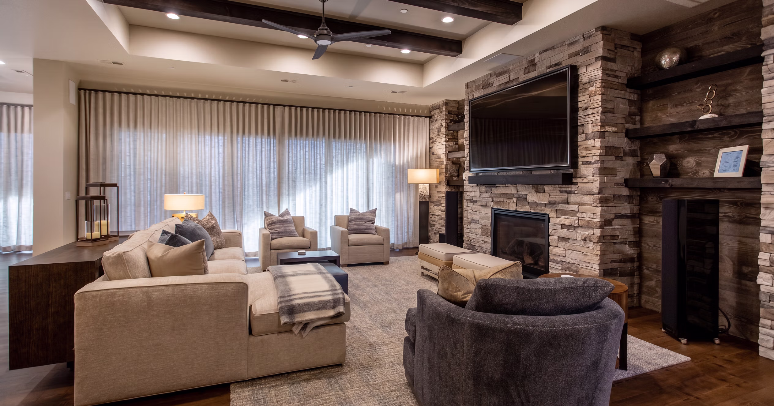 Modern living room with beige sectional sofa, stone fireplace, large TV, and wooden ceiling beams.