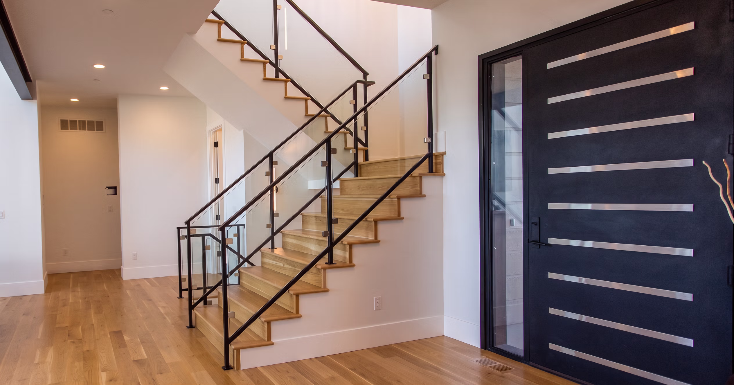 Modern entryway with wooden stairs, glass and metal railing, and a large black door with horizontal metal strips.