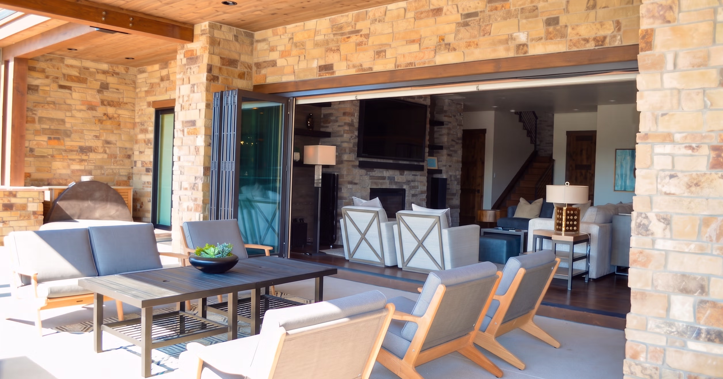 Outdoor patio with stone walls, wooden ceiling, and gray cushioned chairs around a table, opening into a living room.