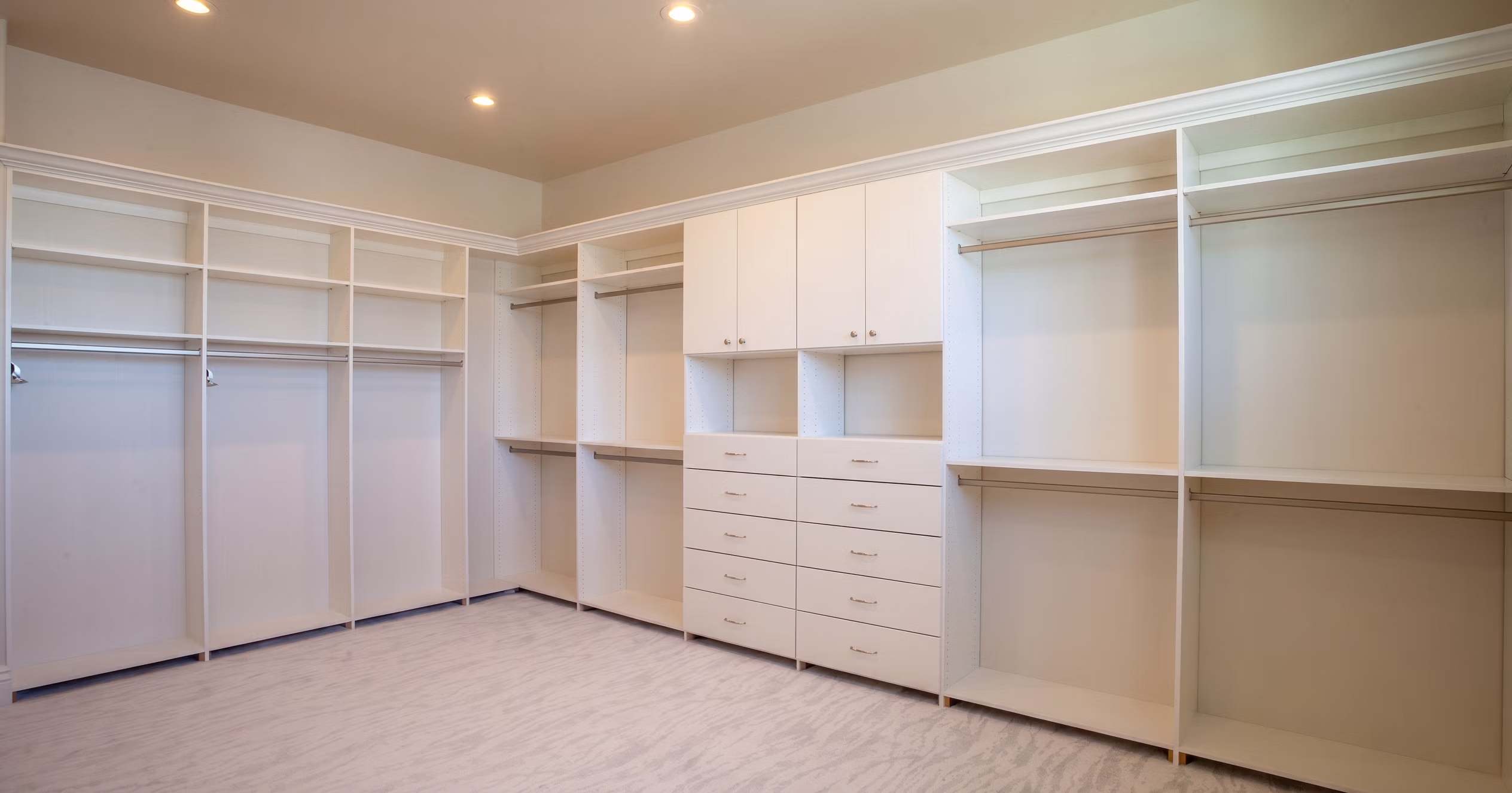 Spacious empty walk-in closet with white built-in shelves, drawers, and hanging rods under ceiling lights.
