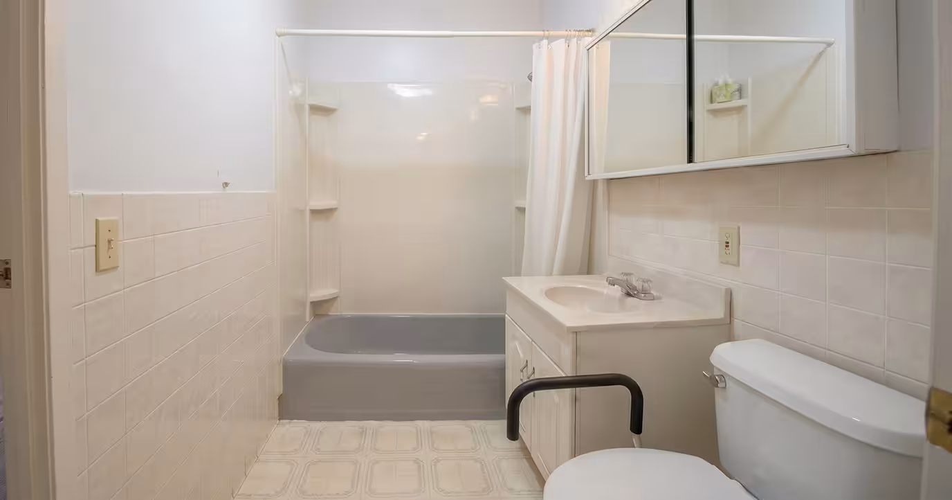 Small bathroom with white tiled walls, gray bathtub, white sink vanity, and toilet with grab bar.
