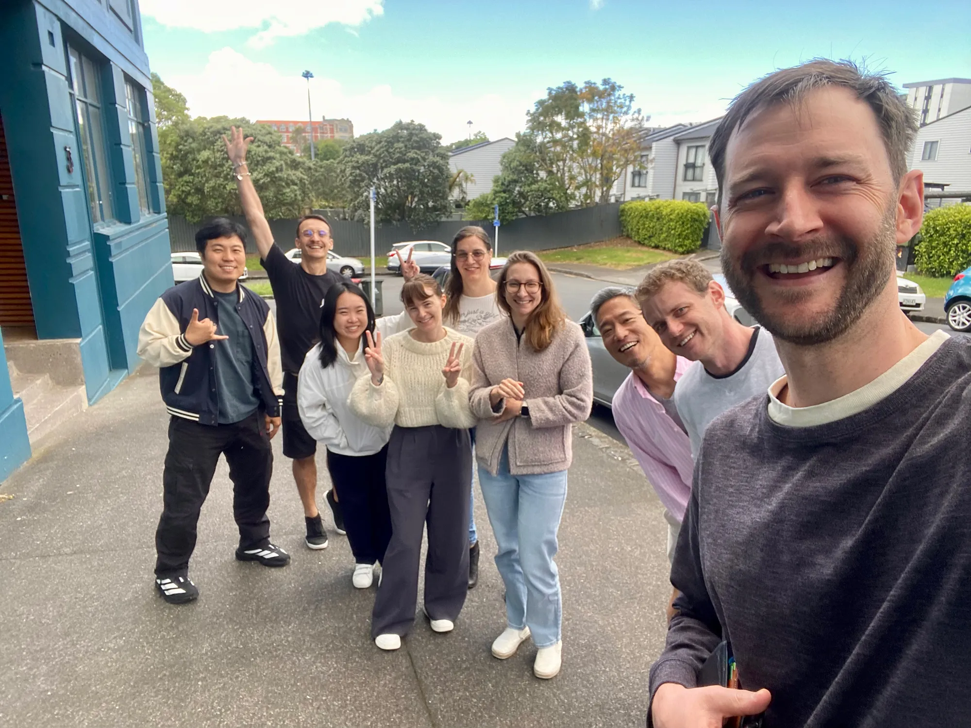 Group of eight people standing outside on a pavement, smiling and posing for a selfie with buildings and greenery in the background.
