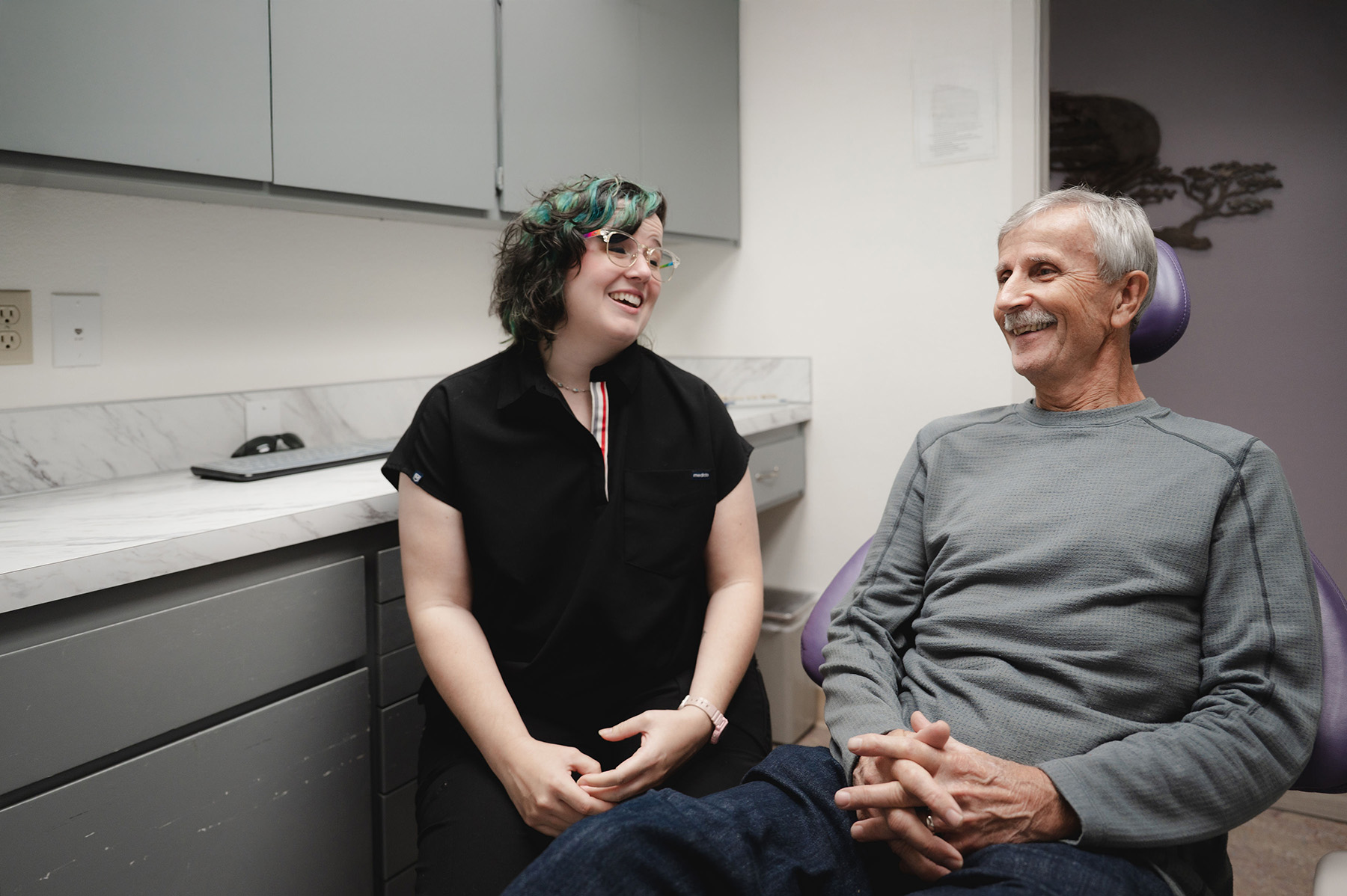 Two men smiling and chatting against a plain white wall, one wearing a red plaid shirt and the other a black t-shirt with colorful tattoo sleeve.