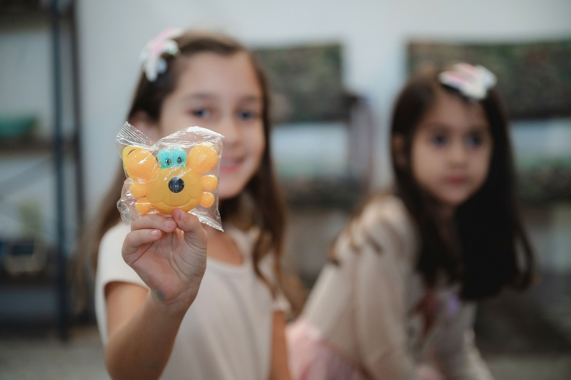 Young girl holding a wrapped orange crab-shaped toy towards the camera with another girl blurred in the background.