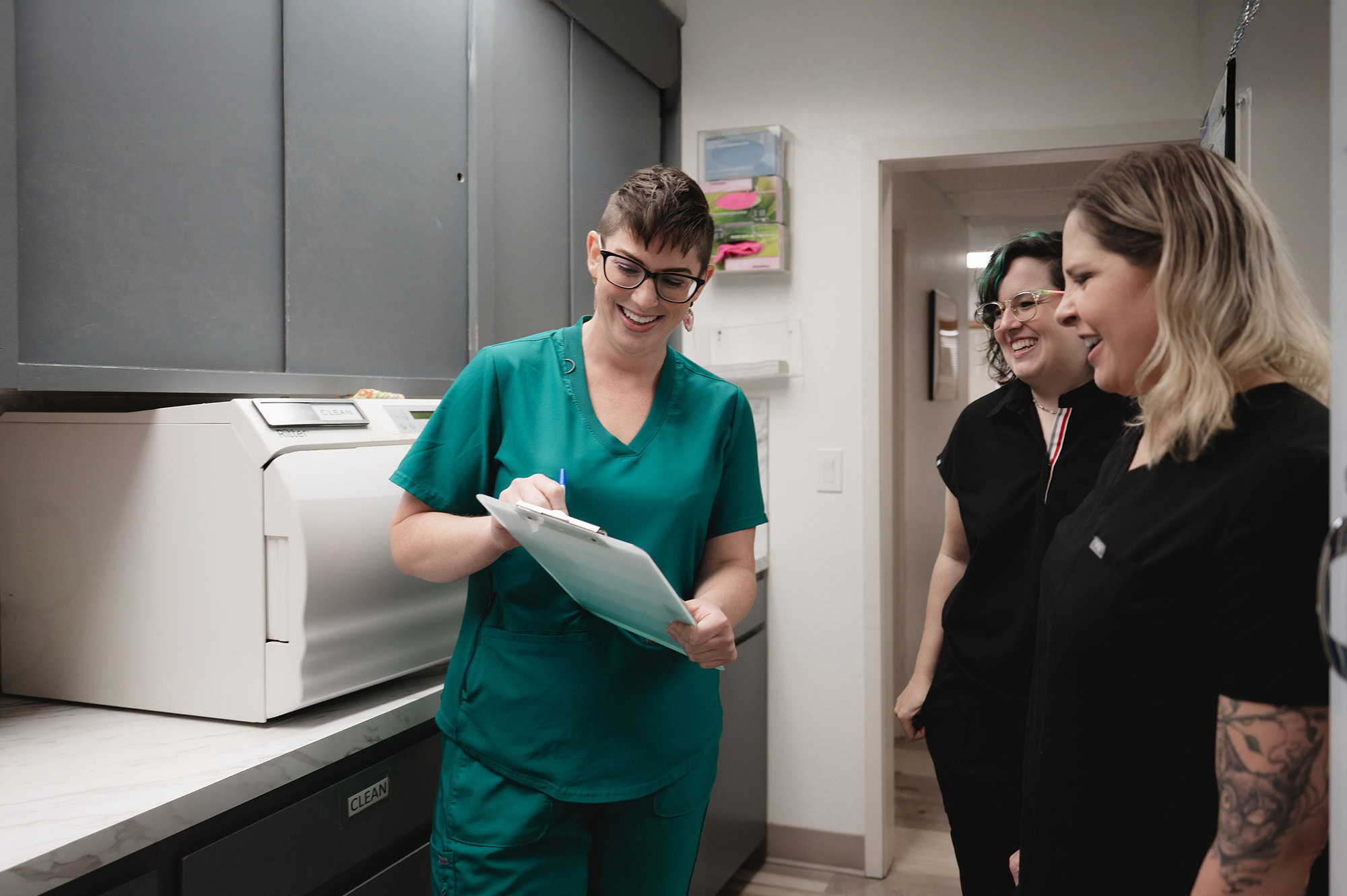 Three smiling medical professionals in scrubs in a medical office, one writing on a clipboard.