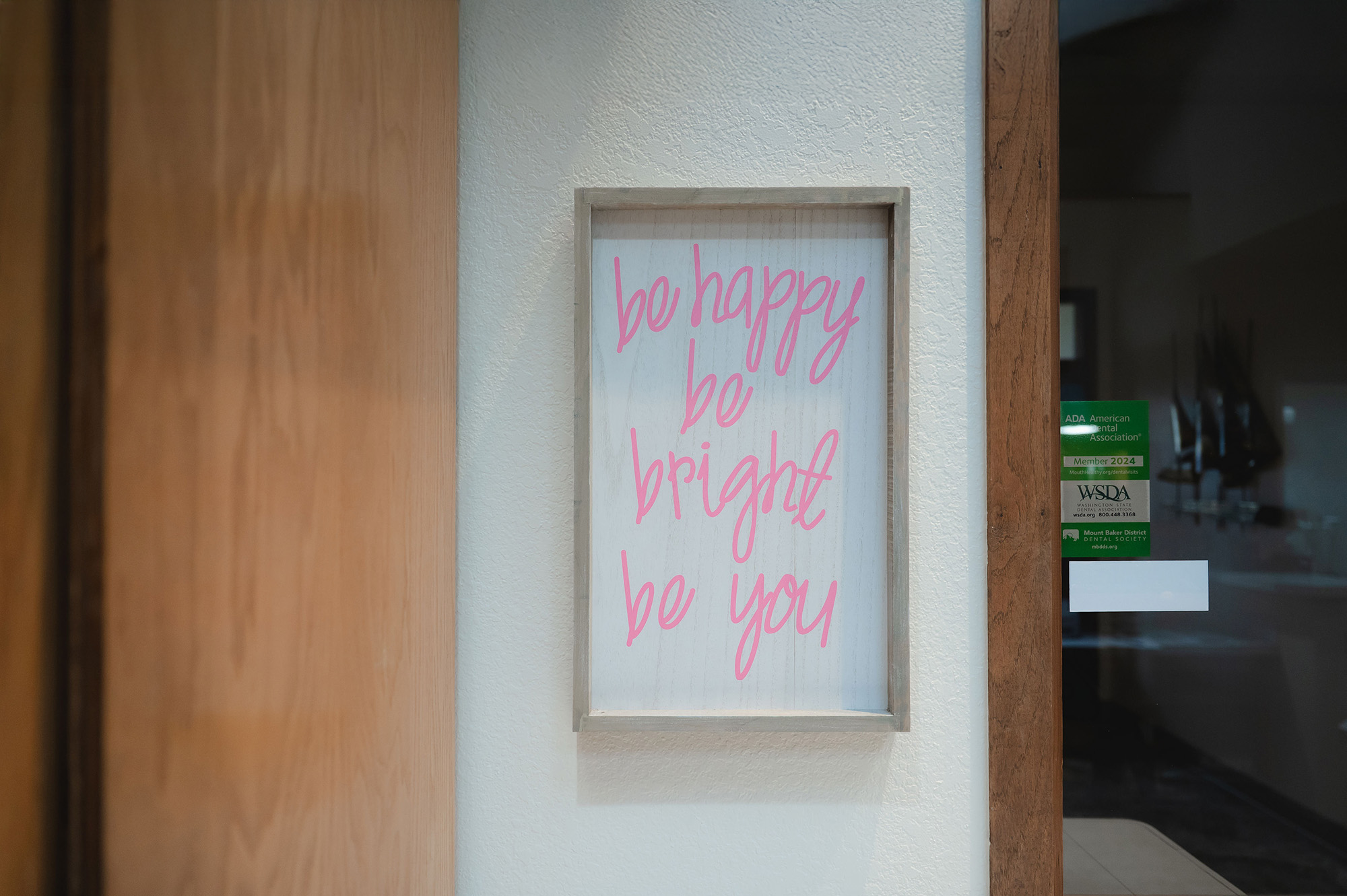 Framed sign on white wall with pink handwritten text saying 'be happy be bright be you'.