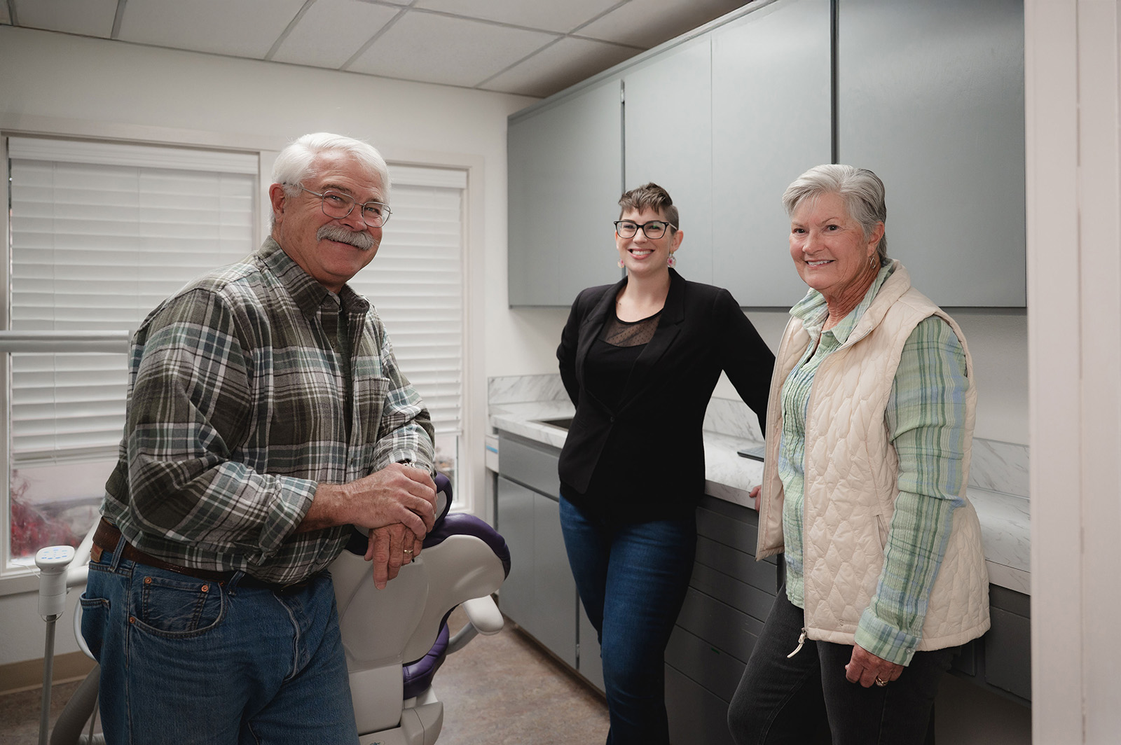 Three smiling adults standing in a dental office near a dental chair and cabinets.