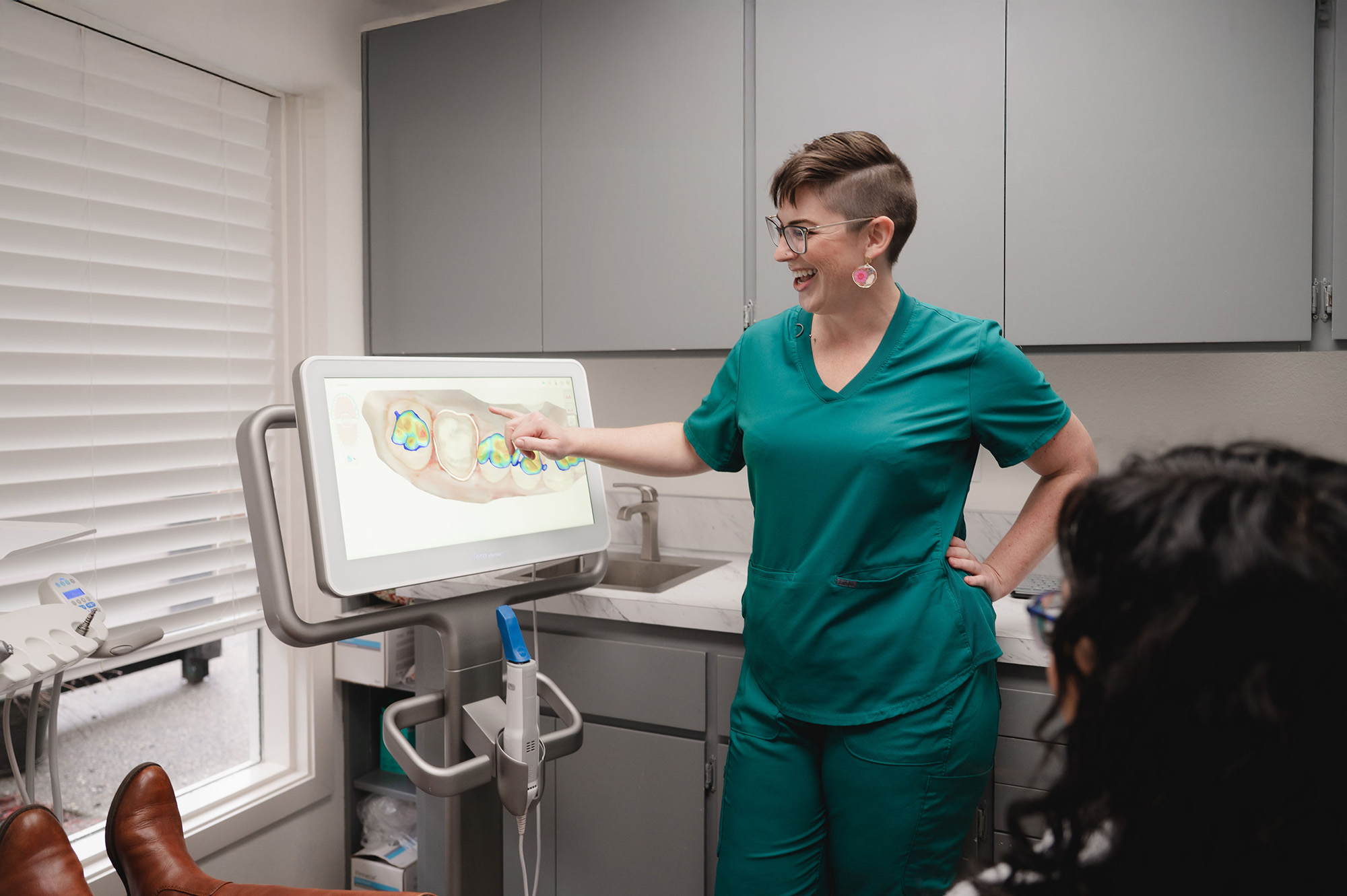 Female dentist in teal scrubs explaining a dental scan on a monitor to a patient in a dental office.