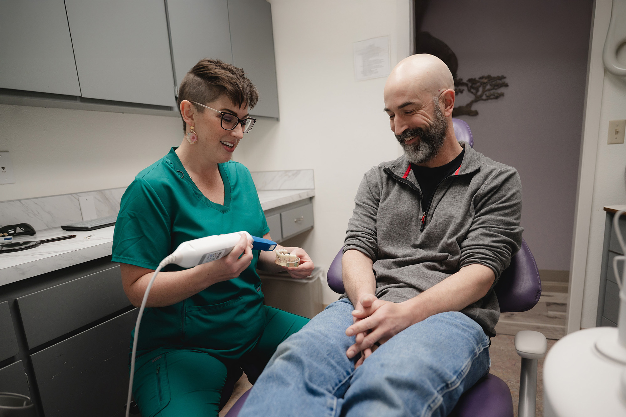 Dentist in green scrubs showing a dental model to a smiling male patient seated in a dental chair.