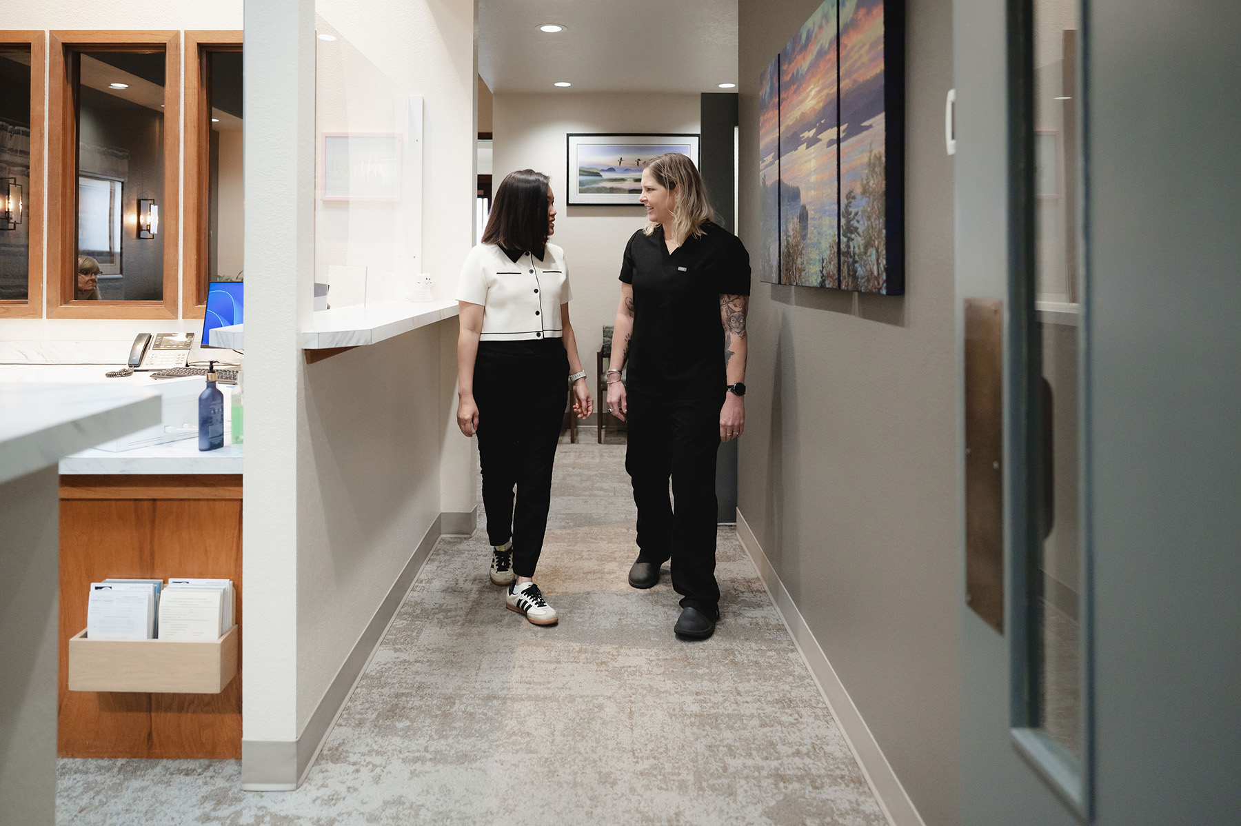 Two women walking down a hallway in a dental office, smiling and conversing, with framed artwork on the walls.