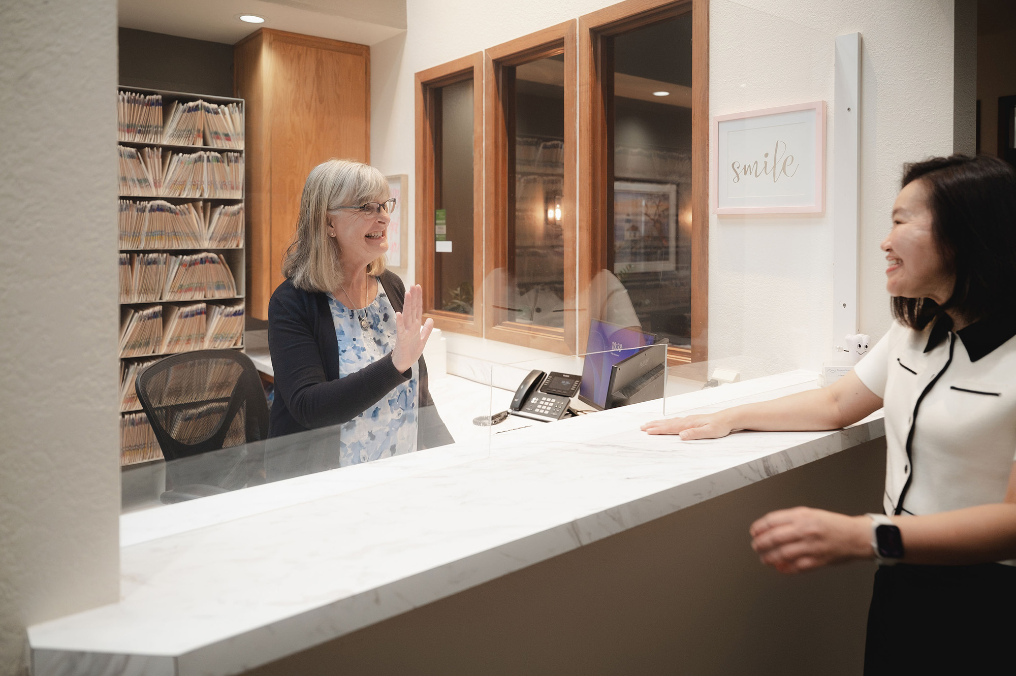 Smiling receptionist behind a desk with a clear protective barrier high-fiving a smiling woman on the other side.