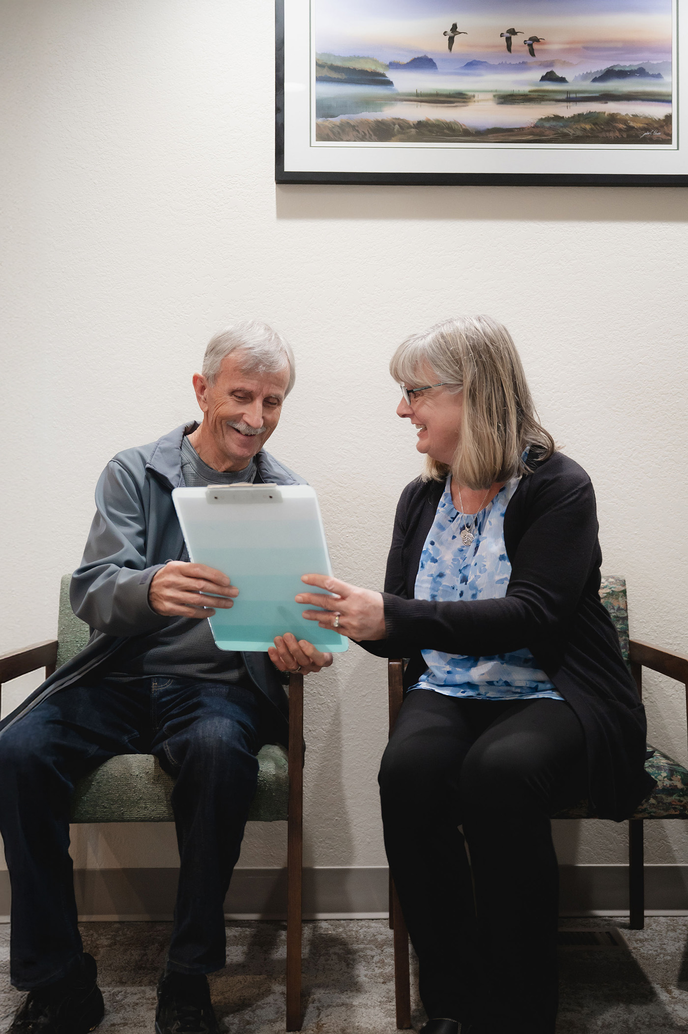 Smiling elderly man and middle-aged woman sitting in chairs reviewing a document on a clipboard.
