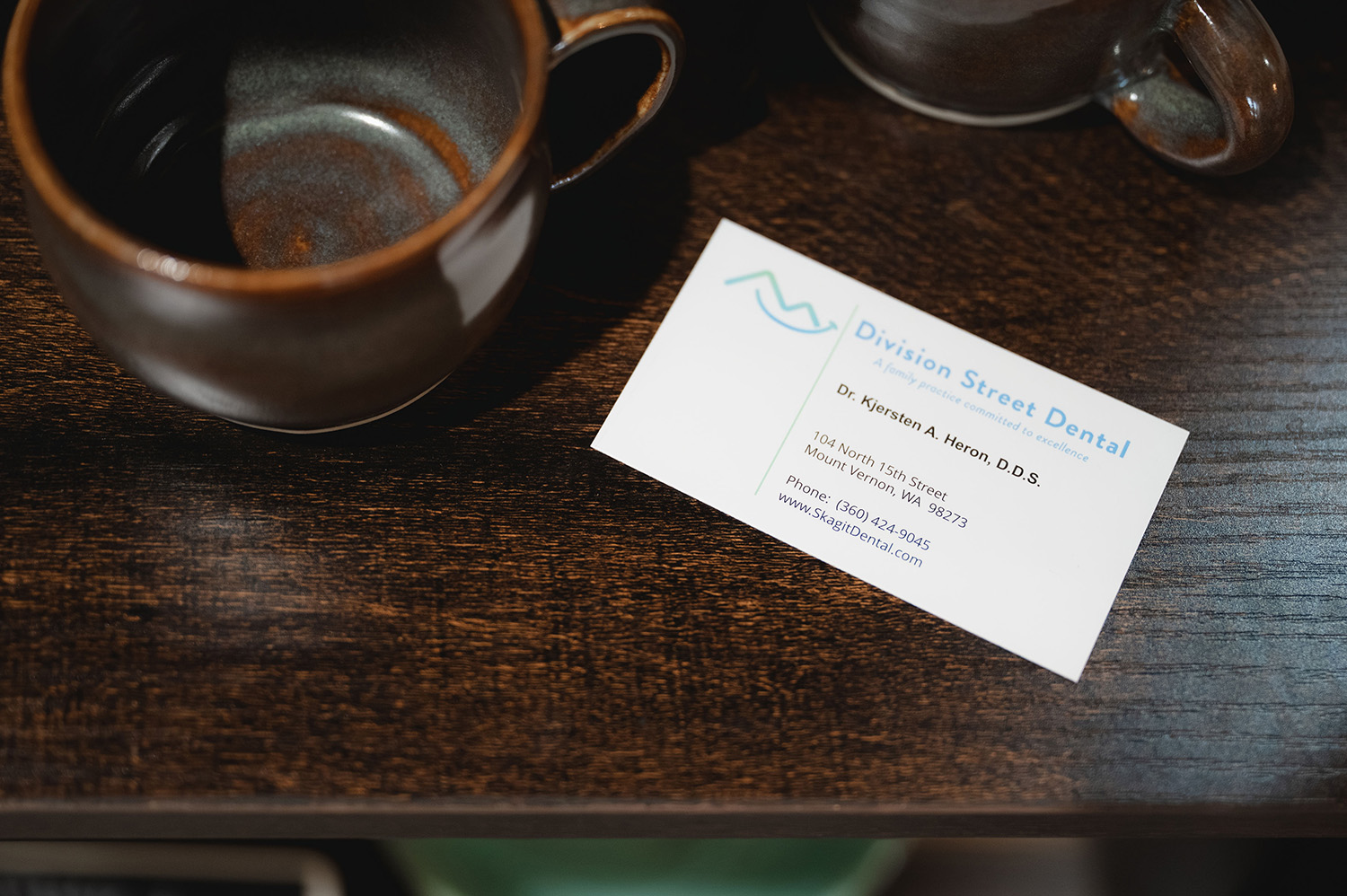 Business card for Division Street Dental with contact details next to two ceramic mugs on a wooden surface.