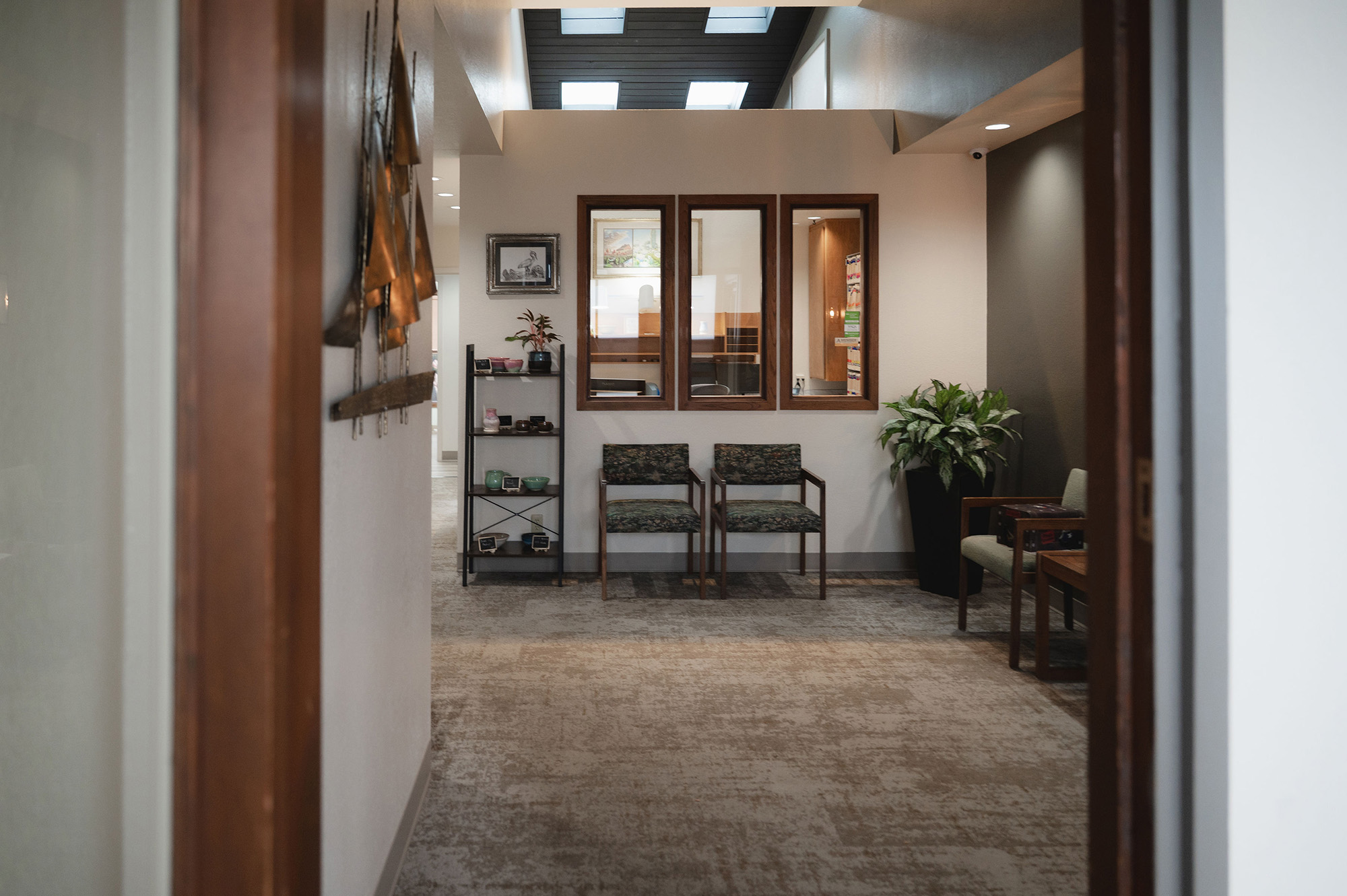 Interior hallway leading to a waiting area with two patterned chairs, a small shelf with decorative items, framed artwork on the wall, and a large potted plant.