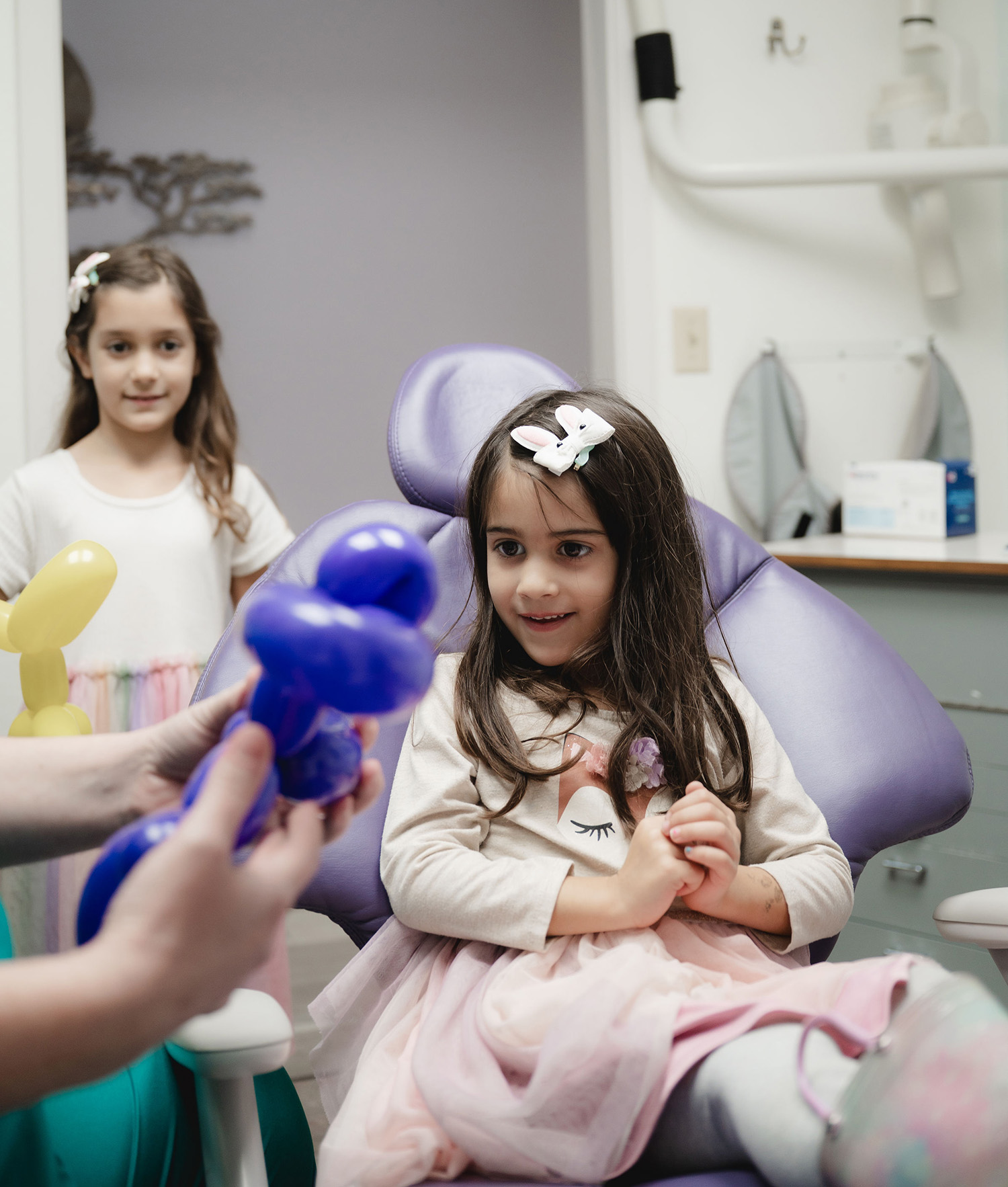 A young girl with a bunny hair clip sits in a purple dental chair looking at a blue balloon animal being made by hands, while another girl with a yellow balloon animal watches nearby.