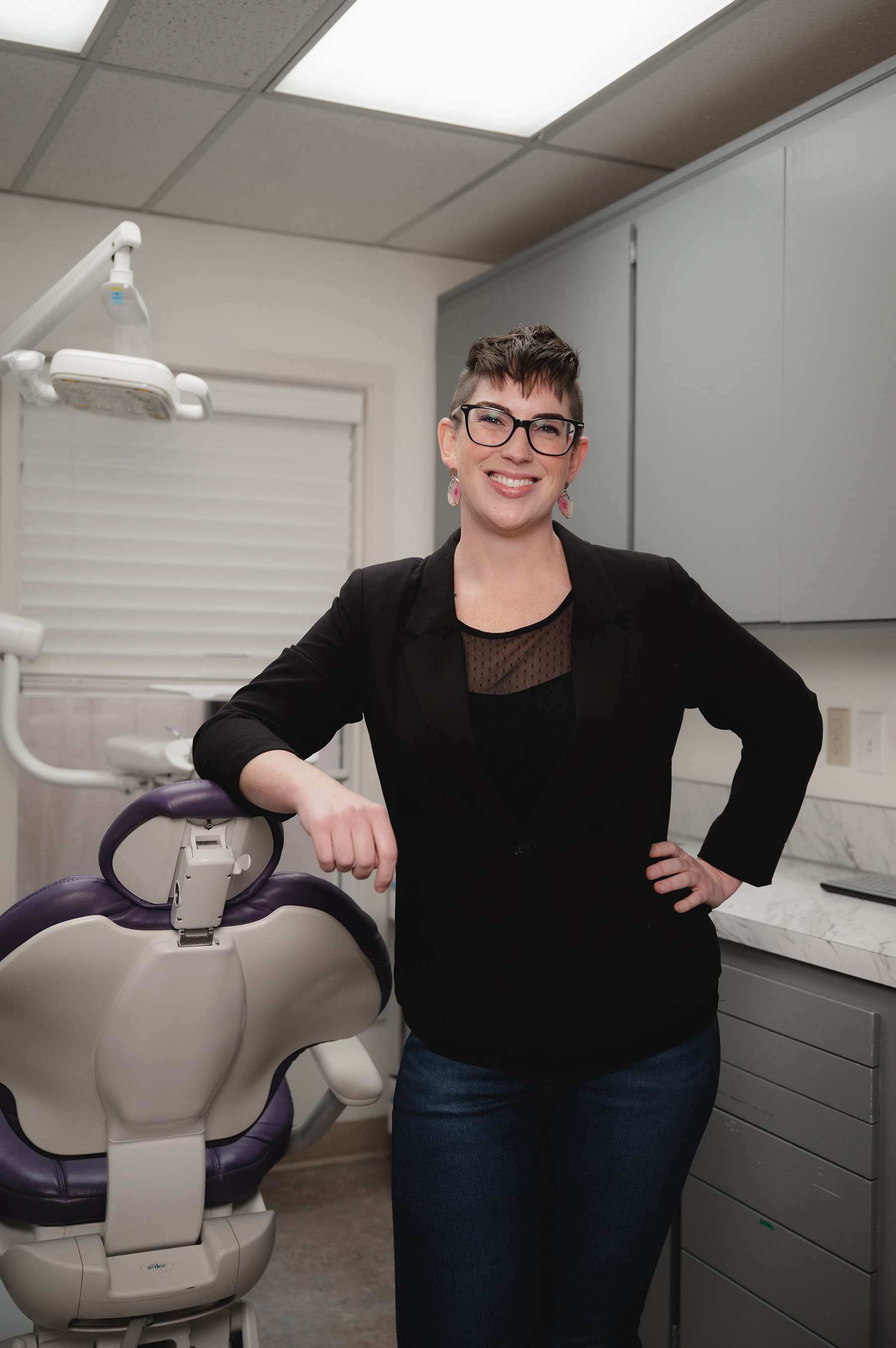 Smiling woman with glasses and earrings leaning on a dental chair in a dental office.