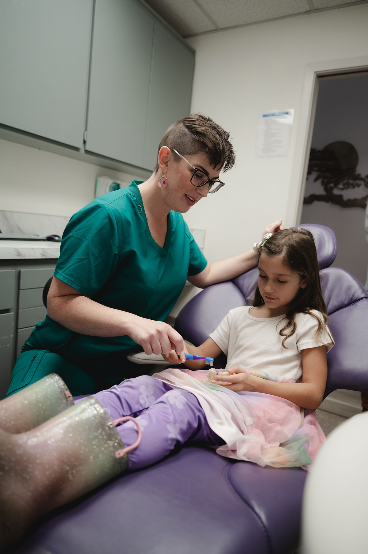Dental professional in green scrubs teaching young girl in purple pants how to brush teeth using a toothbrush and dental model in dental office.